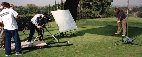 Film crew with camera and reflector filming golfer on green grass with trees in background
