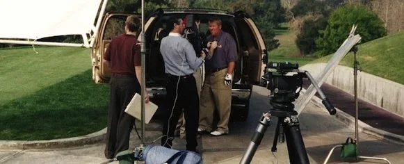 Several men standing beside an open-door vehicle with camera equipment and tripods on a grassy outdoor location.