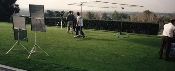 Outdoor film production setup on grass field with lighting reflectors, crew members, and equipment
