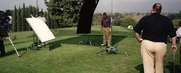 Film or photo production crew setting up lighting equipment on a grassy outdoor field with reflectors and stands