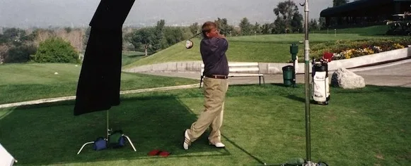 Golfer mid-swing on a practice range with trees and landscaped grounds in background