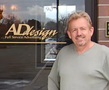 Man in light gray shirt standing in front of storefront with Ad Design signage