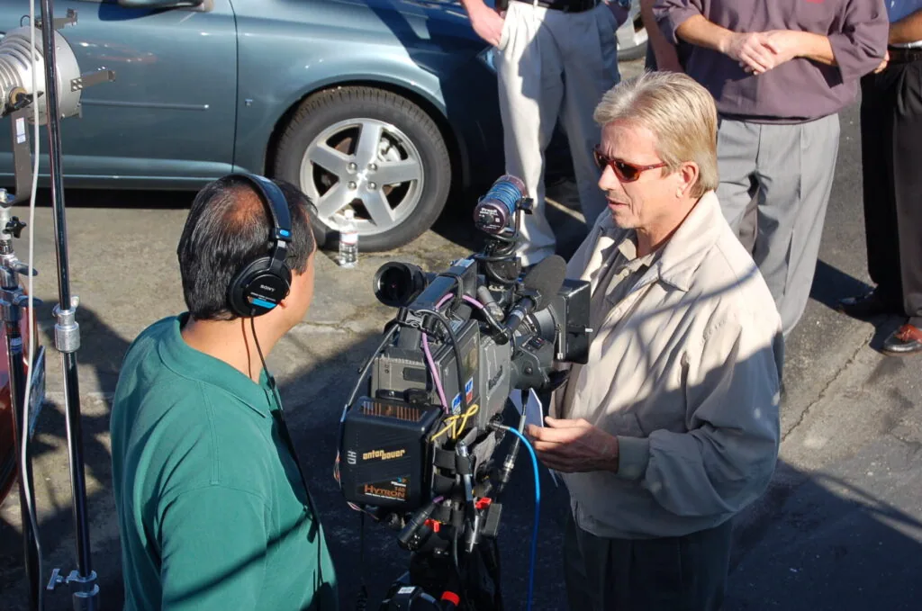 Man in sunglasses and tan jacket speaking with person wearing headphones and green shirt at outdoor event near parked vehicles and video camera equipment