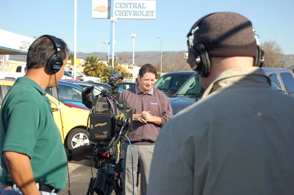 Two men wearing headphones interview a man in front of parked cars at a Chevrolet dealership