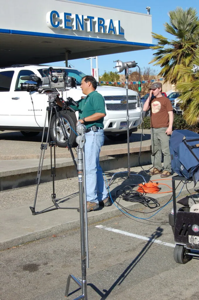 Two people operating a professional video camera on a tripod at an outdoor vehicle dealership lot with "CENTRAL" signage visible.