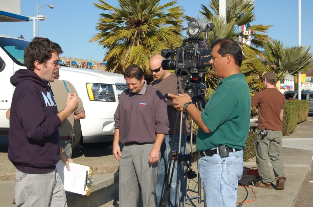 Film production crew operating camera and conducting interview on location with palm trees and commercial building in background