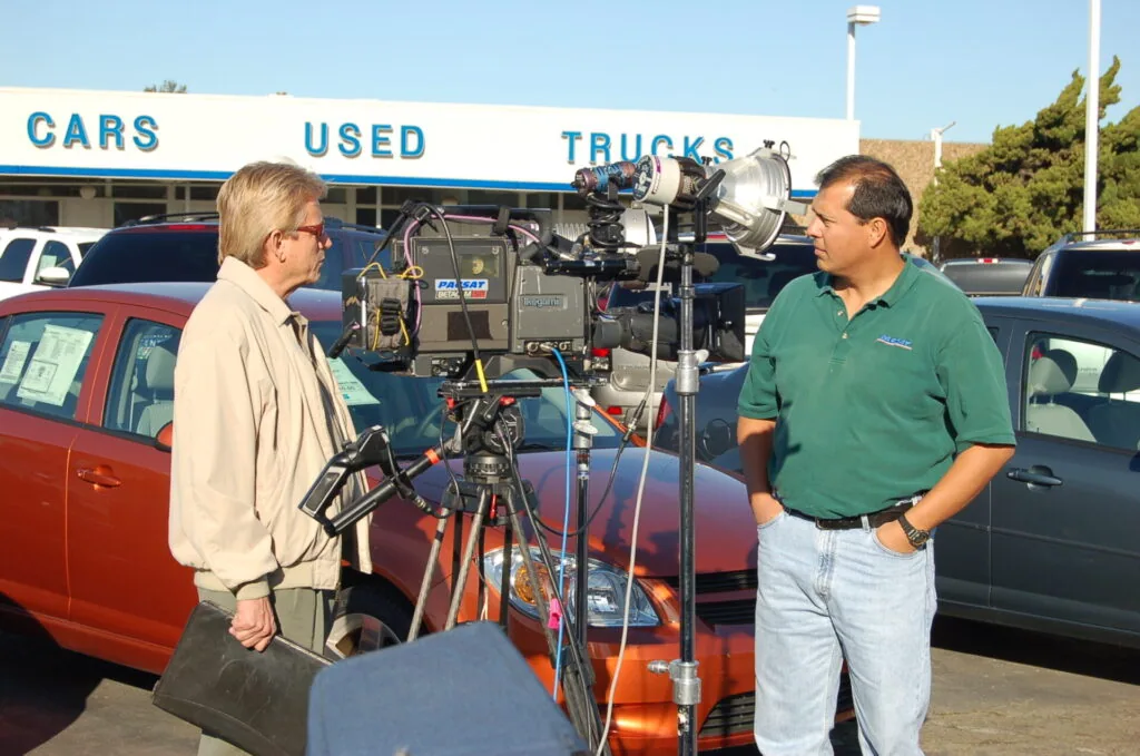 Two men standing between film cameras and lighting equipment at a used cars and trucks dealership lot