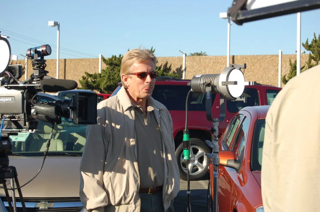 Man in sunglasses and tan jacket standing on film set with professional lighting equipment and red vehicles in background
