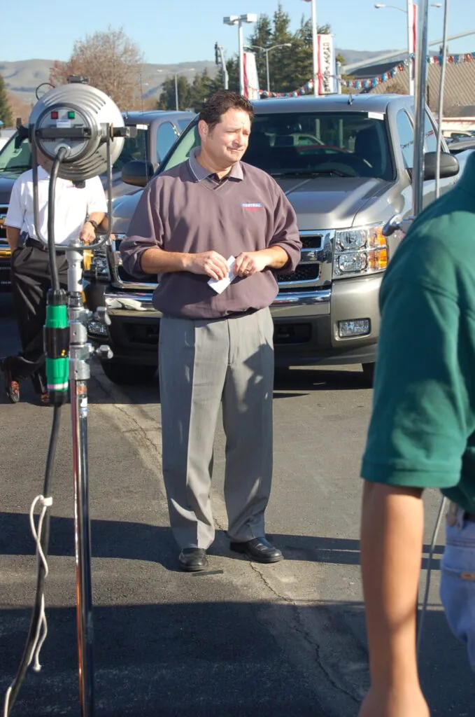 Man in brown polo shirt standing at outdoor event with bicycle and camera equipment in parking lot