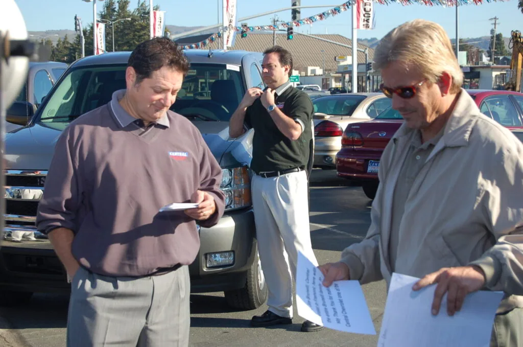 Three men in a car lot examining documents, with vehicles and promotional banners visible in the background