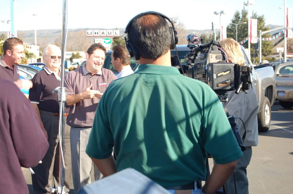 Film crew recording interview with multiple people in parking lot near U-Haul sign and vehicles