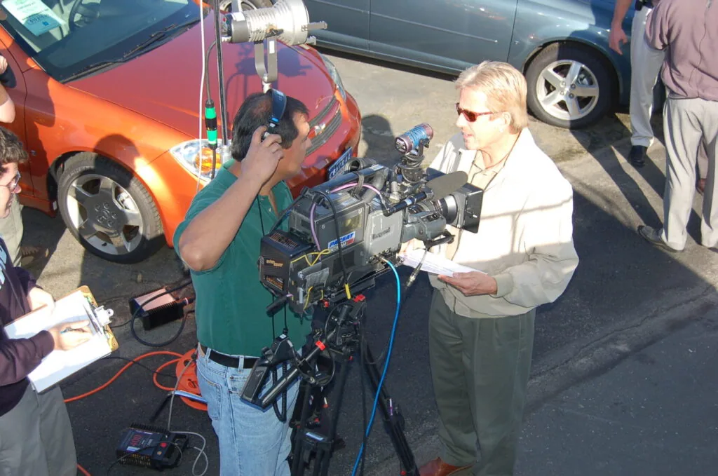Cameraman films interview in parking lot with orange car and crew members present